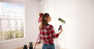 Close up. Side view of pretty joyful Caucasian woman alone painting room in new house in green color. Beautiful female renovating home painting wall using roller brush redesigning apartment