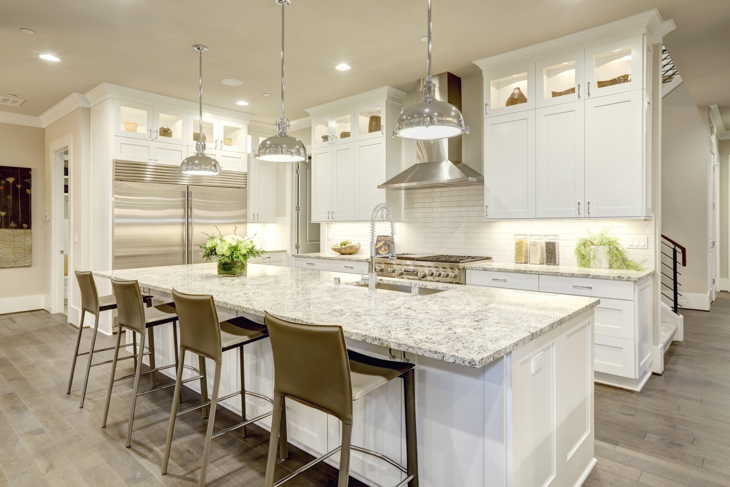 White kitchen design features large bar style kitchen island with granite countertop illuminated by modern pendant lights. Stainless steel appliances framed by white shaker cabinets . Northwest, USA